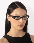 A woman with long, straight, black hair embodies vintage charm in her For Art's Sake® Cigar black cat-eye glasses and sleeveless top. She glances slightly to the side against a plain, light background.