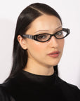 A woman with long, straight black hair is wearing the "Cigar" cat-eye glasses from For Art's Sake®, featuring thick black frames that lend her a vintage charm. She is dressed in a black turtleneck top and is gazing directly at the camera against a plain white background.