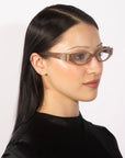 A person with long black hair is shown from a side angle, wearing For Art's Sake® Cigar beige frame eyeglasses and a black top. The background is plain white, emphasizing the person's sleek, minimalist look with a touch of vintage charm.