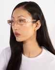 A person with long, straight hair and wearing transparent pink For Art's Sake® Cigar frames looks to the side. They are dressed in a white top, exuding a serene expression. The background is plain white, adding a contemporary edge to the scene.