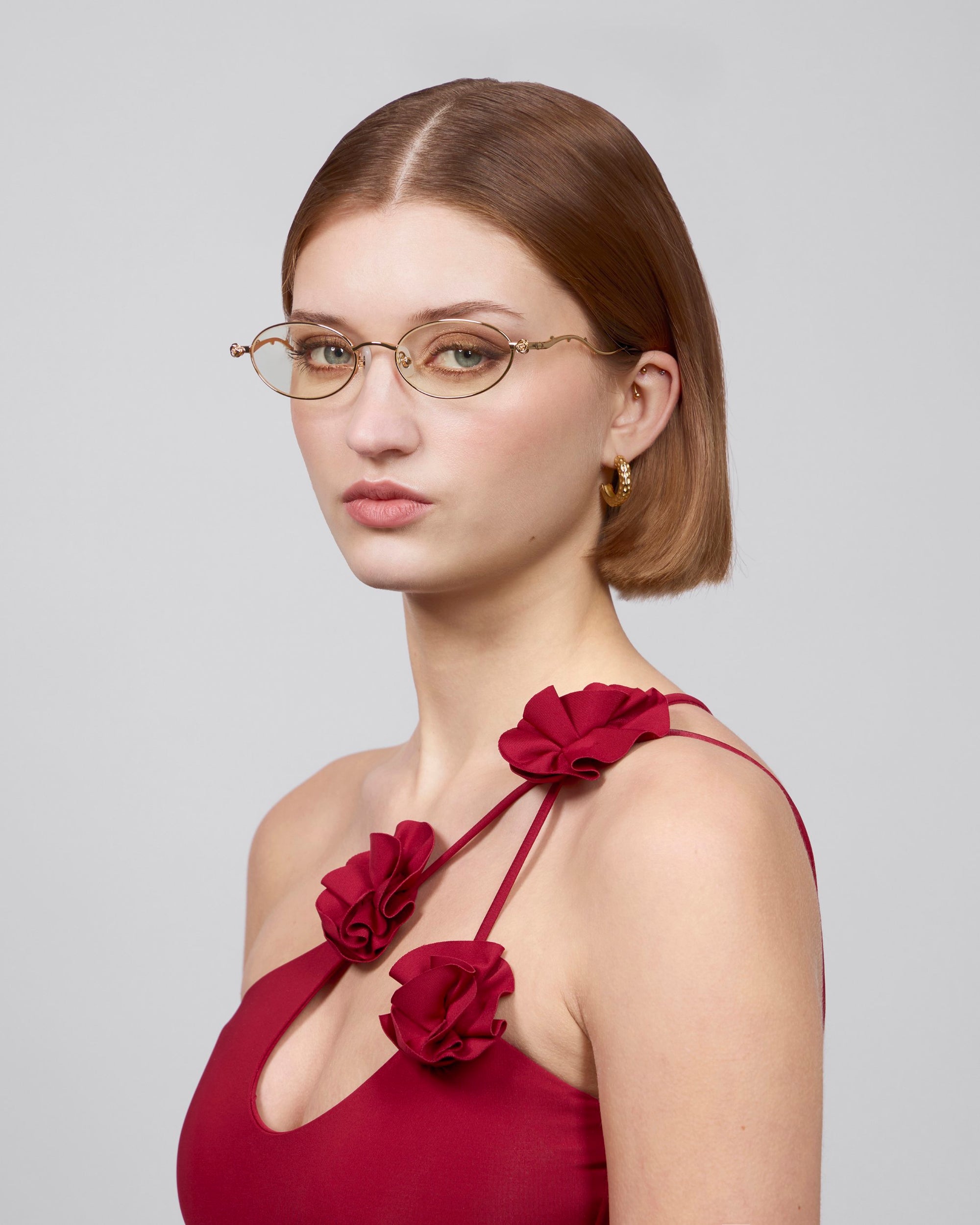 A person with short brown hair wearing gold-rimmed glasses and a sleeveless red "Thorn" dress from For Art's Sake®. They accessorize with gold hoop earrings and pose against a plain background, embodying an elegant, statement masterpiece.