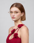 A person with short brown hair wearing gold-rimmed glasses and a sleeveless red "Thorn" dress from For Art's Sake®. They accessorize with gold hoop earrings and pose against a plain background, embodying an elegant, statement masterpiece.