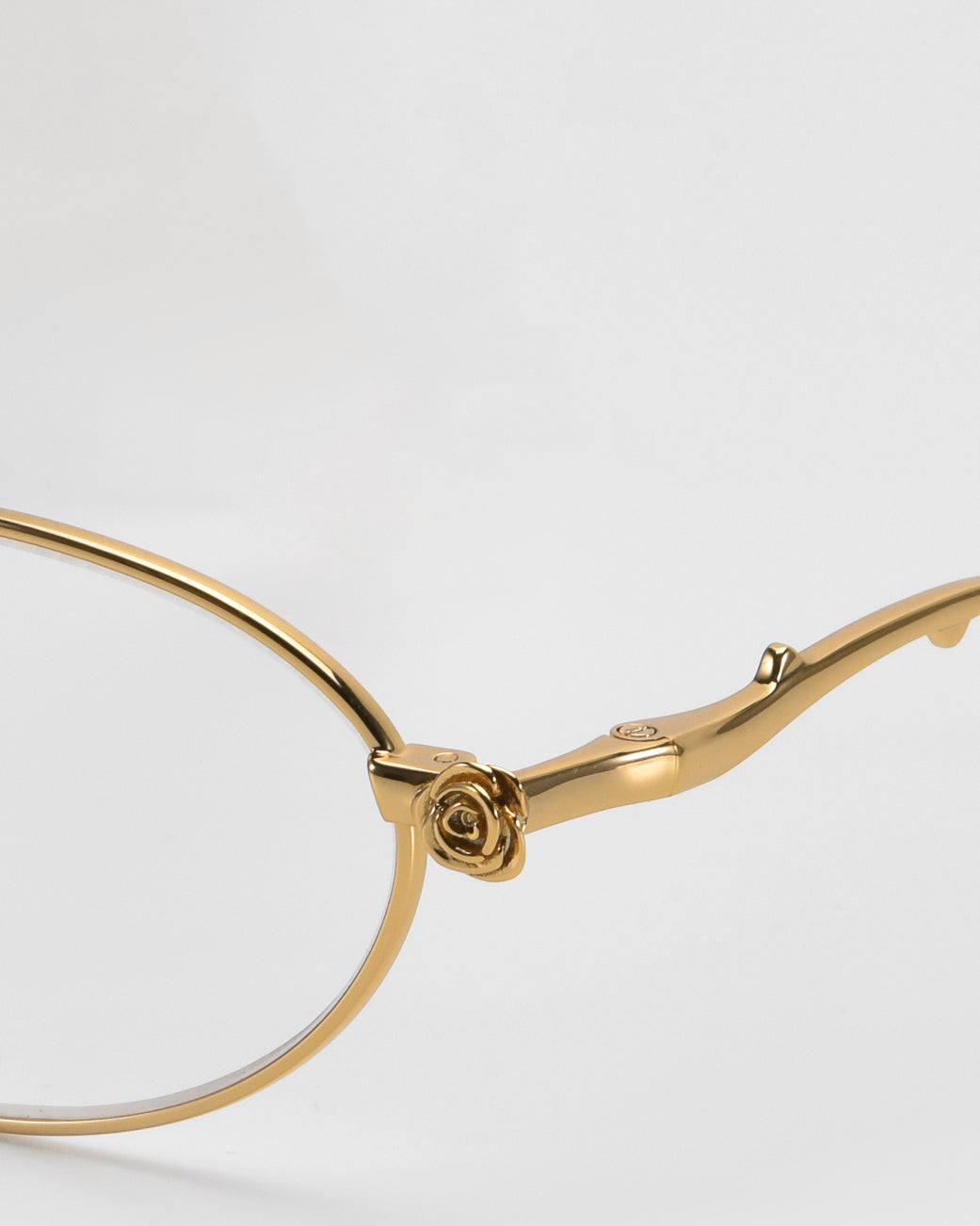 This close-up of the gold Thorn eyeglass frames by For Art's Sake® features a delicate rose detail on the hinge. The polished finish adds elegance, making it a true statement piece. The neutral background complements its exquisite oval silhouette beautifully.