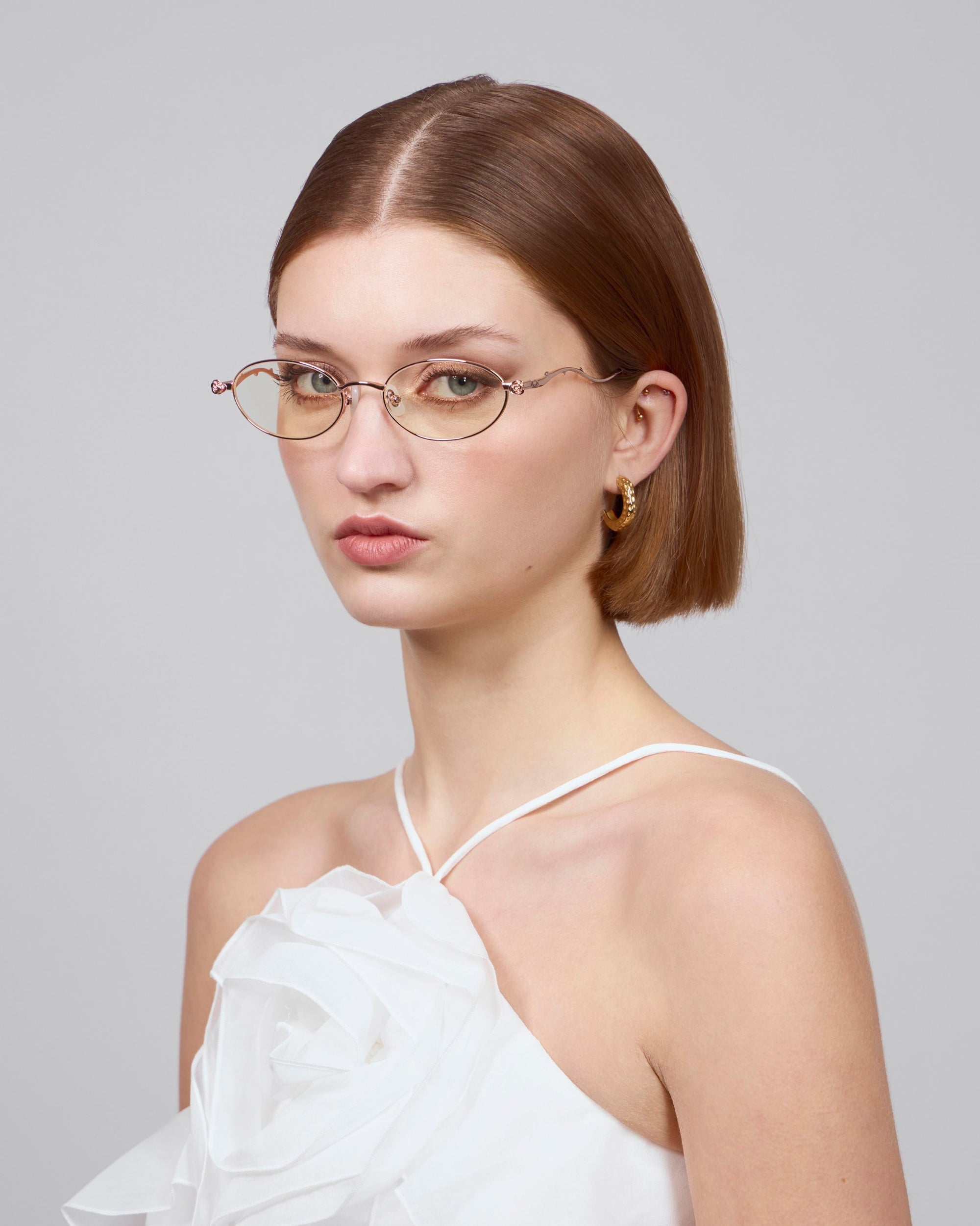 A woman with short brown hair wears thin-framed, small round glasses and gold hoop earrings. She dons a white top with a large fabric flower detail from the Rose Garden collection. The background is plain gray. Glasses: Thorn by For Art's Sake®.