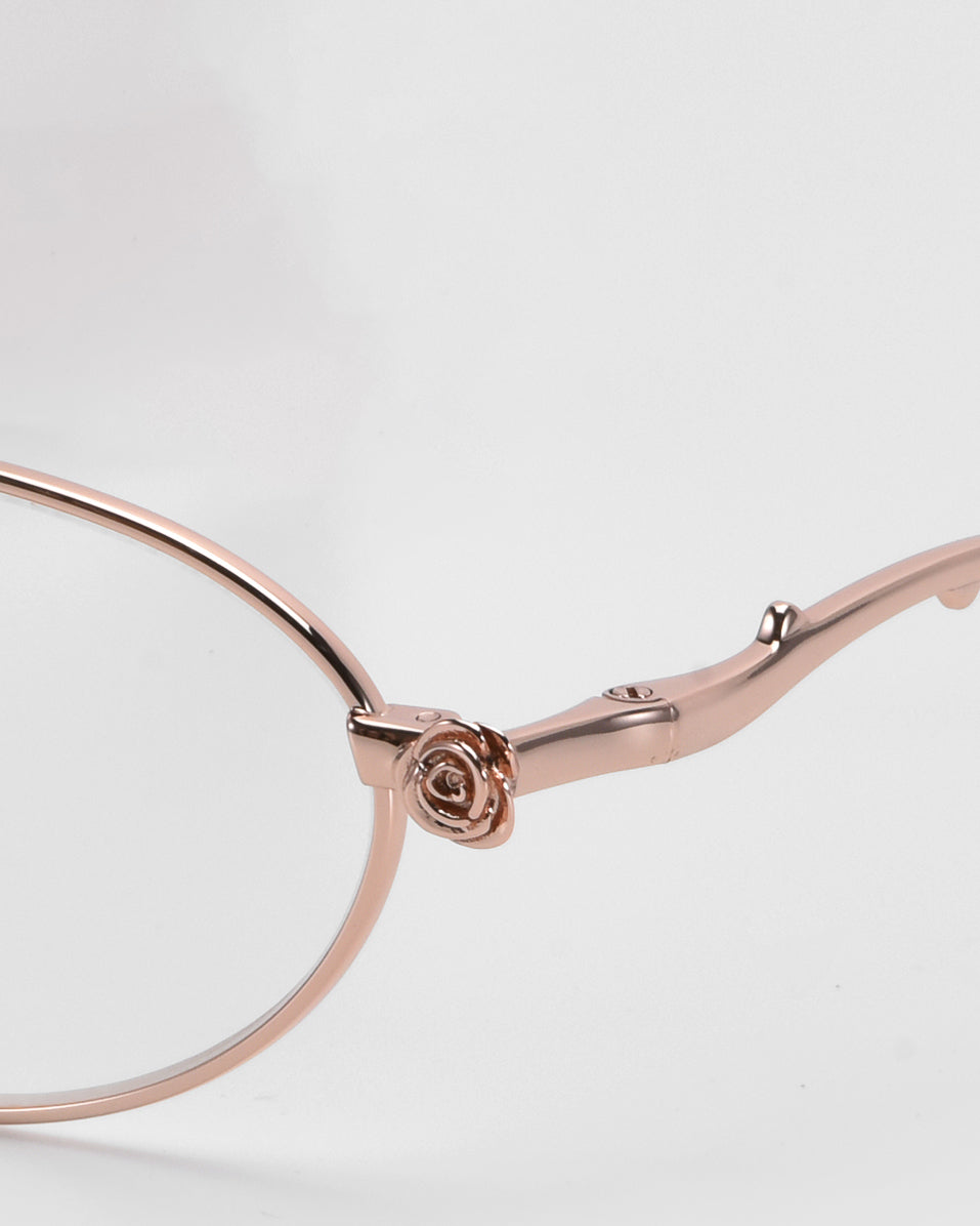 Close-up of For Art's Sake® Thorn eyeglasses, showcasing a delicate rose detail on the temple arm. The modern oval silhouette frame is in a soft rose gold color with a smooth, elegant finish against a neutral background.