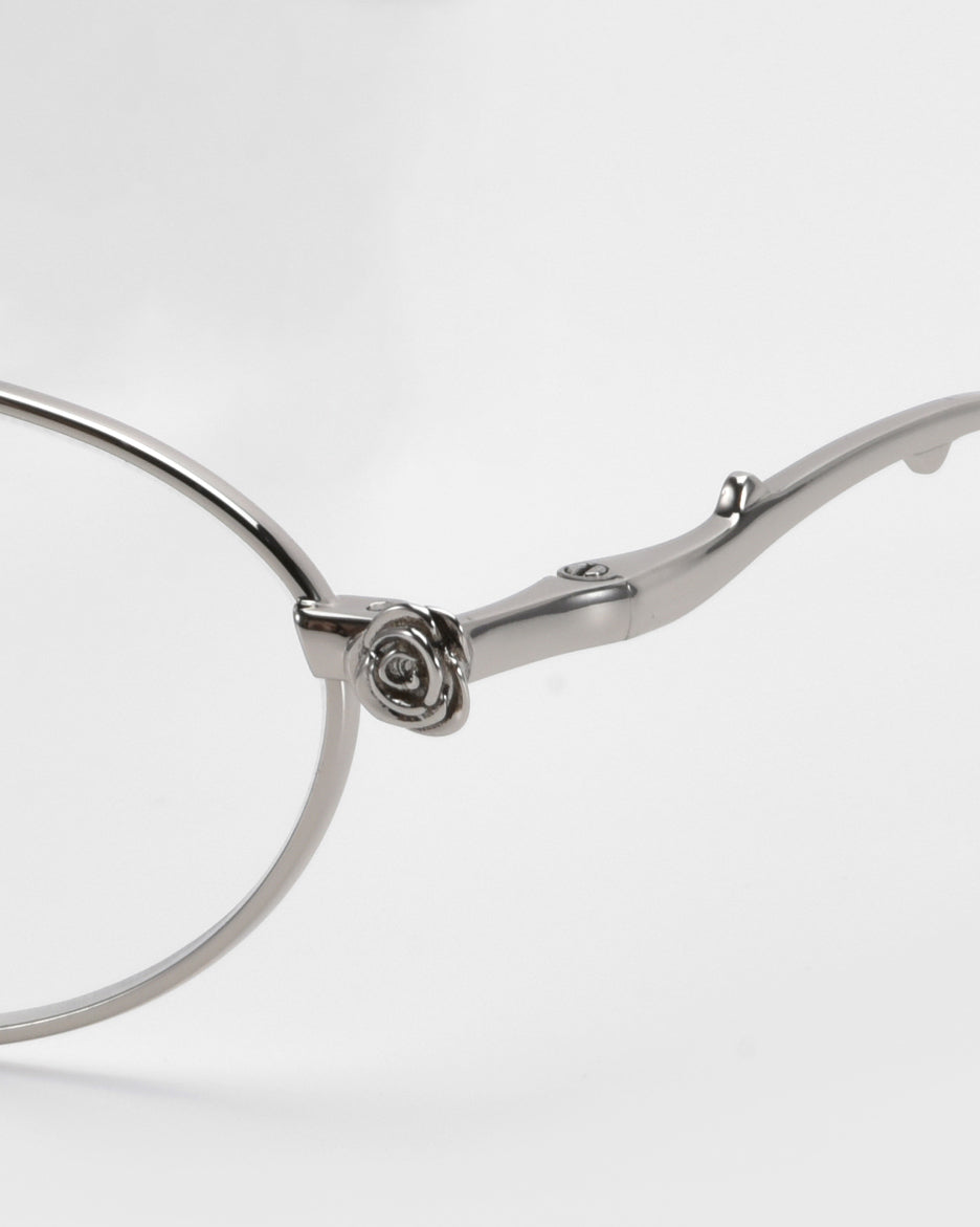 Close-up of For Art's Sake® Thorn eyeglasses. The silver metal frames feature floral detailing at the temple and hinge, leading to round lenses. This statement piece from the Rose Garden collection brilliantly stands out against a plain white background.