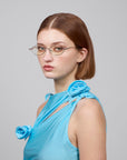 A person with short brown hair, wearing For Art's Sake® optical glasses and a light blue sleeveless top with floral designs, looks slightly to the side against a plain gray background, showcasing their unique sense of artistic expression through the "Thorn" eyewear.