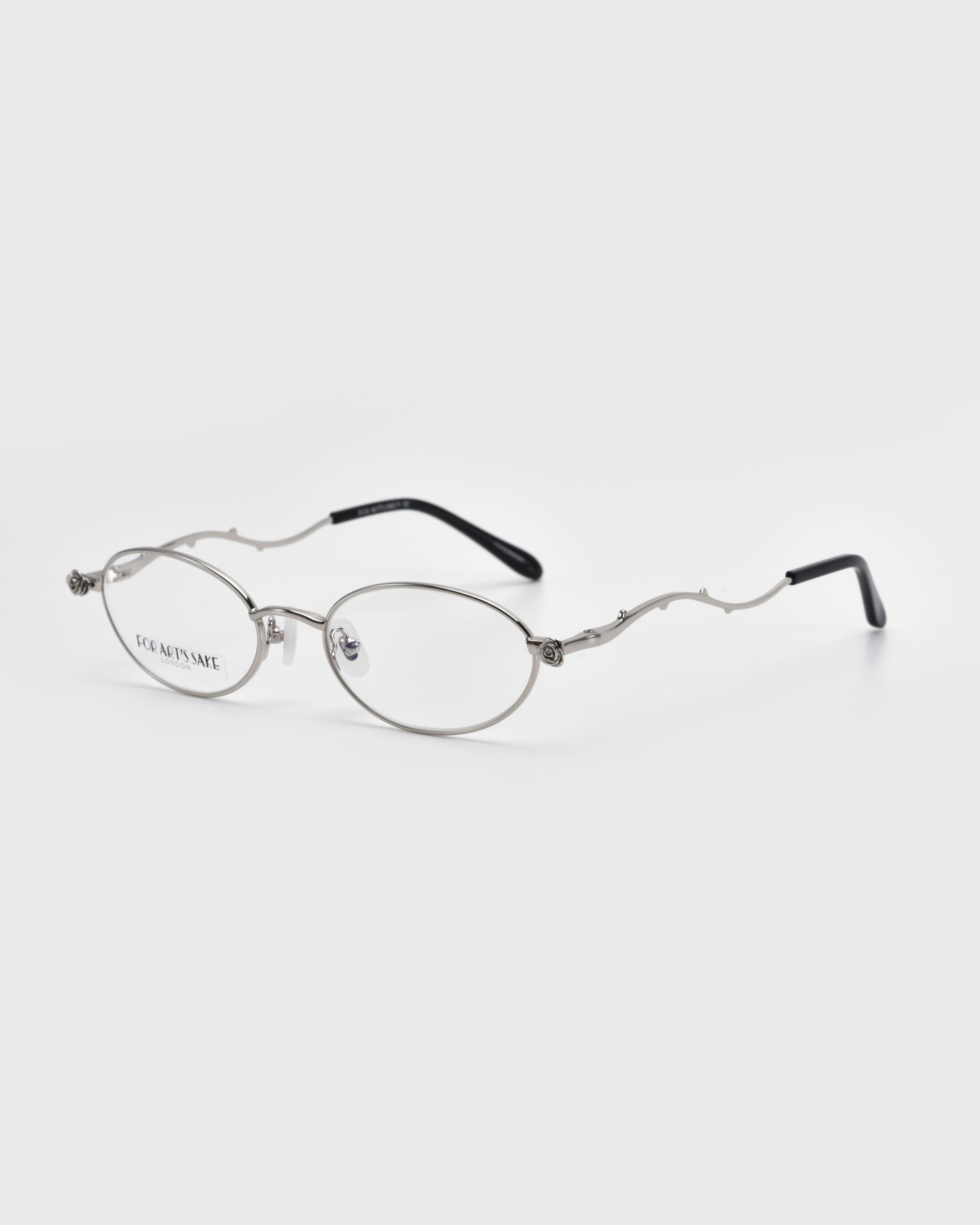 For Art's Sake® presents the Thorn eyeglasses, part of the Rose Garden collection, with silver frames featuring oval lenses and thorn-inspired temple arms. The sleek metallic look is enhanced by black end tips and clear lenses against a white background.