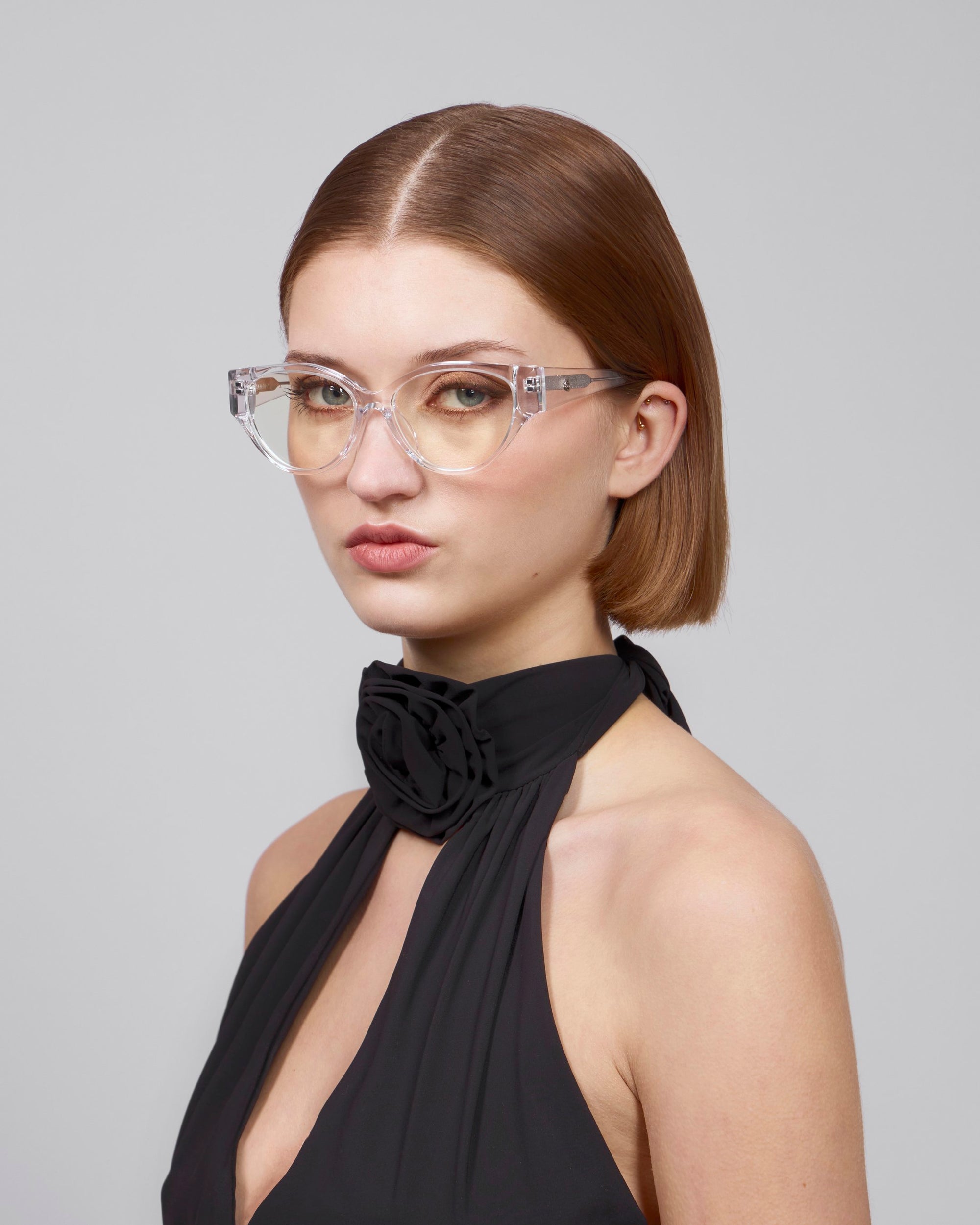 A woman with short brown hair exudes femininity in transparent cat-eye frames, Rosalia I by For Art's Sake®, paired with a black halter top adorned with floral detail. Against a plain gray backdrop, she epitomizes the Rose Garden Collection.