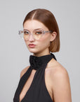 A woman with short brown hair exudes femininity in transparent cat-eye frames, Rosalia I by For Art's Sake®, paired with a black halter top adorned with floral detail. Against a plain gray backdrop, she epitomizes the Rose Garden Collection.