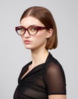 A person with short brown hair wears Rosalia I red cat-eye optical frames by For Art's Sake® and a black sheer short-sleeved top. They are turned slightly to the side, looking directly at the camera against a plain grey background, exuding femininity.