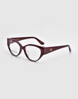 The Rosalia I eyeglasses from For Art's Sake® are part of the Rose Garden Collection, blending femininity and elegance with purple oval lenses, tapered arms with a gold emblem on a light gray background. The clear lenses are ideal for prescriptions.