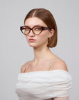 A woman with short brown hair exudes femininity, wearing large cat-eye optical frames from For Art's Sake® Rosalia I and gold hoop earrings. Her off-the-shoulder white top against a plain gray background embodies the elegance of the Rose Garden Collection.