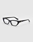 The Rosalia II by For Art's Sake® features stylish black rectangular eyeglasses from the Rose Garden Collection with a glossy finish. A small golden emblem on the temple adds femininity, resting elegantly on a light gray surface.