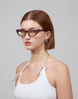 A person with short brown hair wears the Rosalia II cat-eye optical frame from For Art's Sake®, paired with silver hoop earrings and a white tank top with thin straps. Against a plain gray background, they exude femininity while gazing directly at the camera.