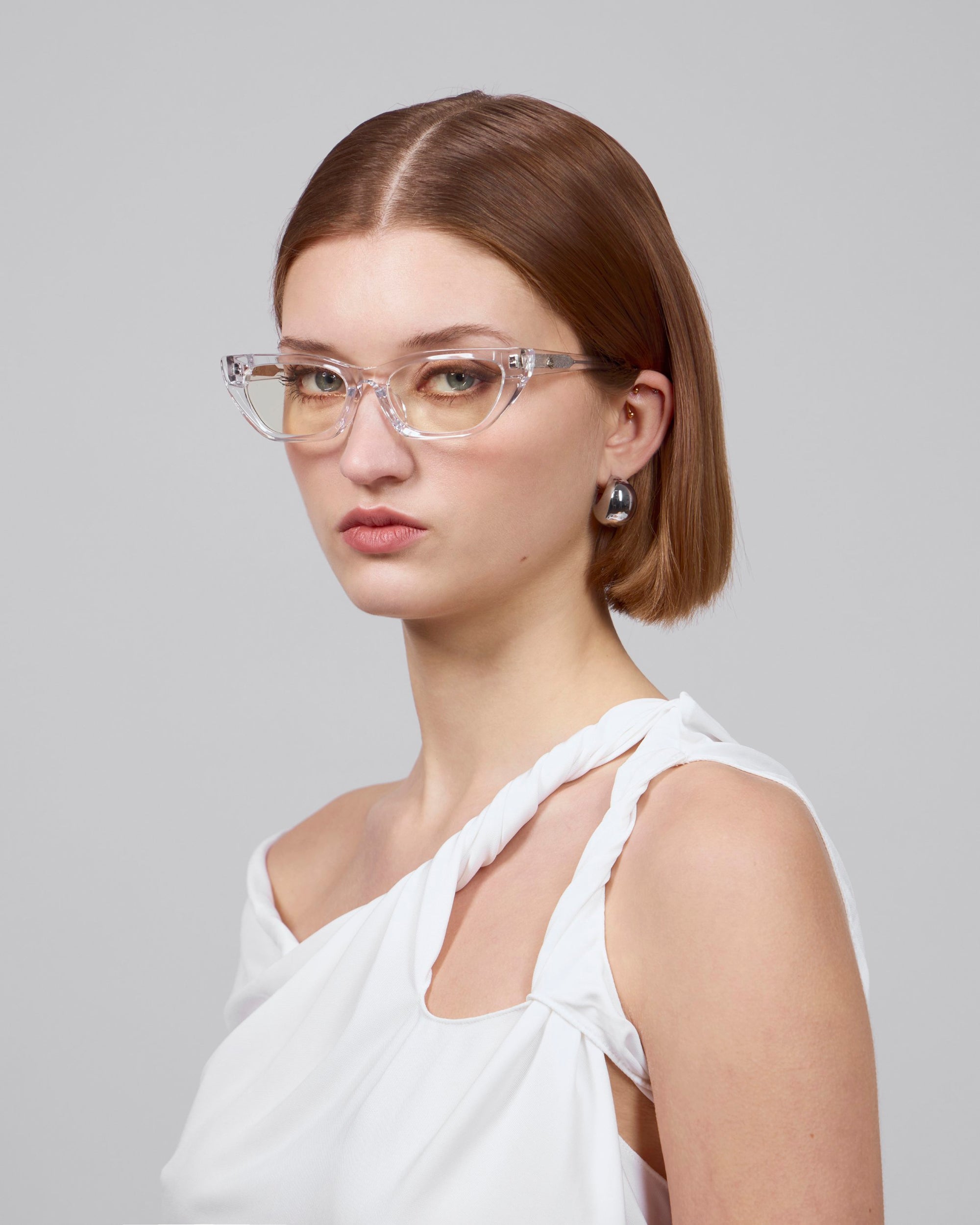 A short-haired individual with brown locks, transparent glasses, and pearl earrings exudes elegance while gazing into the camera, adorned in For Art's Sake® Rosalia II—a white, one-shoulder top—against a plain gray backdrop.