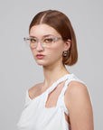 A short-haired individual with brown locks, transparent glasses, and pearl earrings exudes elegance while gazing into the camera, adorned in For Art's Sake® Rosalia II—a white, one-shoulder top—against a plain gray backdrop.