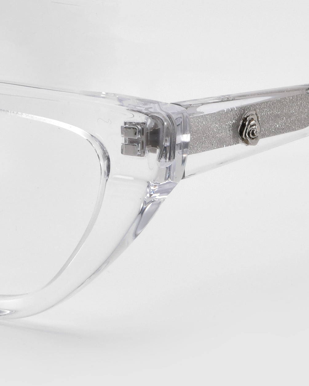 Close-up of the Rosalia II cat-eye optical frame by For Art's Sake® from the Rose Garden Collection. The clear frames exude femininity, featuring a silver temple arm with textured finish and circular detail, elegantly displayed against a soft white background.