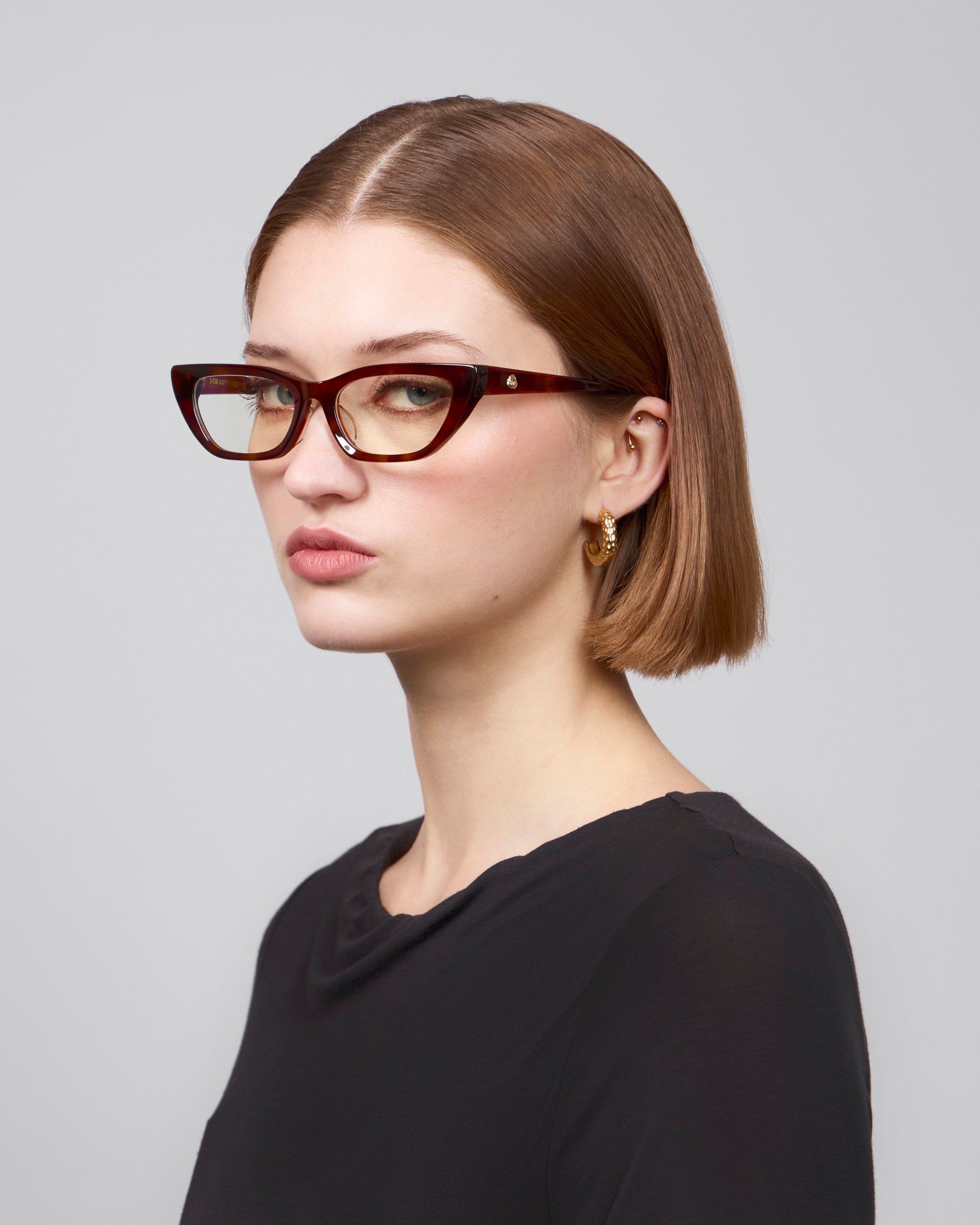 A person with short brown hair wears For Art's Sake® Rosalia II cat-eye glasses and a black top, exuding femininity with gold hoop earrings. They gaze slightly to the side against a neutral background.