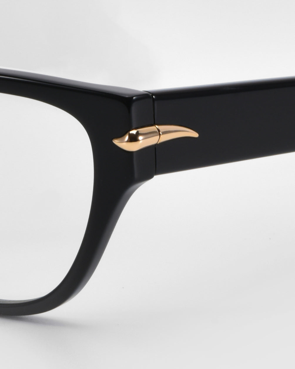 Close-up of For Art's Sake® Eros eyeglass frames in black, highlighted by a gold wing-like decoration on the arm hinge, evocative of cat-eye acetate design. The background is plain and white.