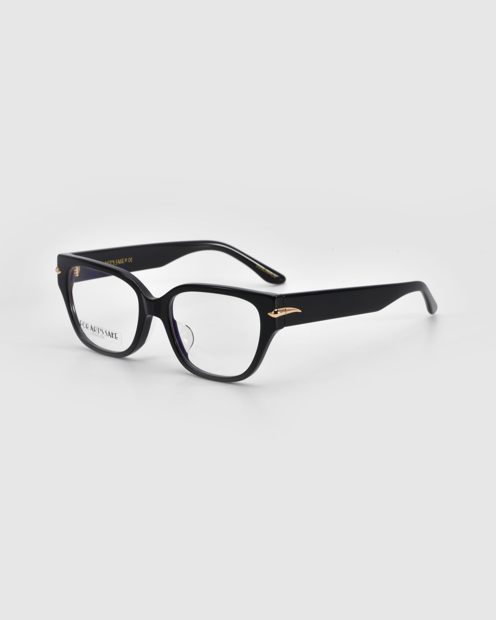 The For Art's Sake® Eros eyeglasses rest on a light gray surface, featuring black rectangular acetate frames with a subtle cat-eye shape and elegant golden accents near the hinges.