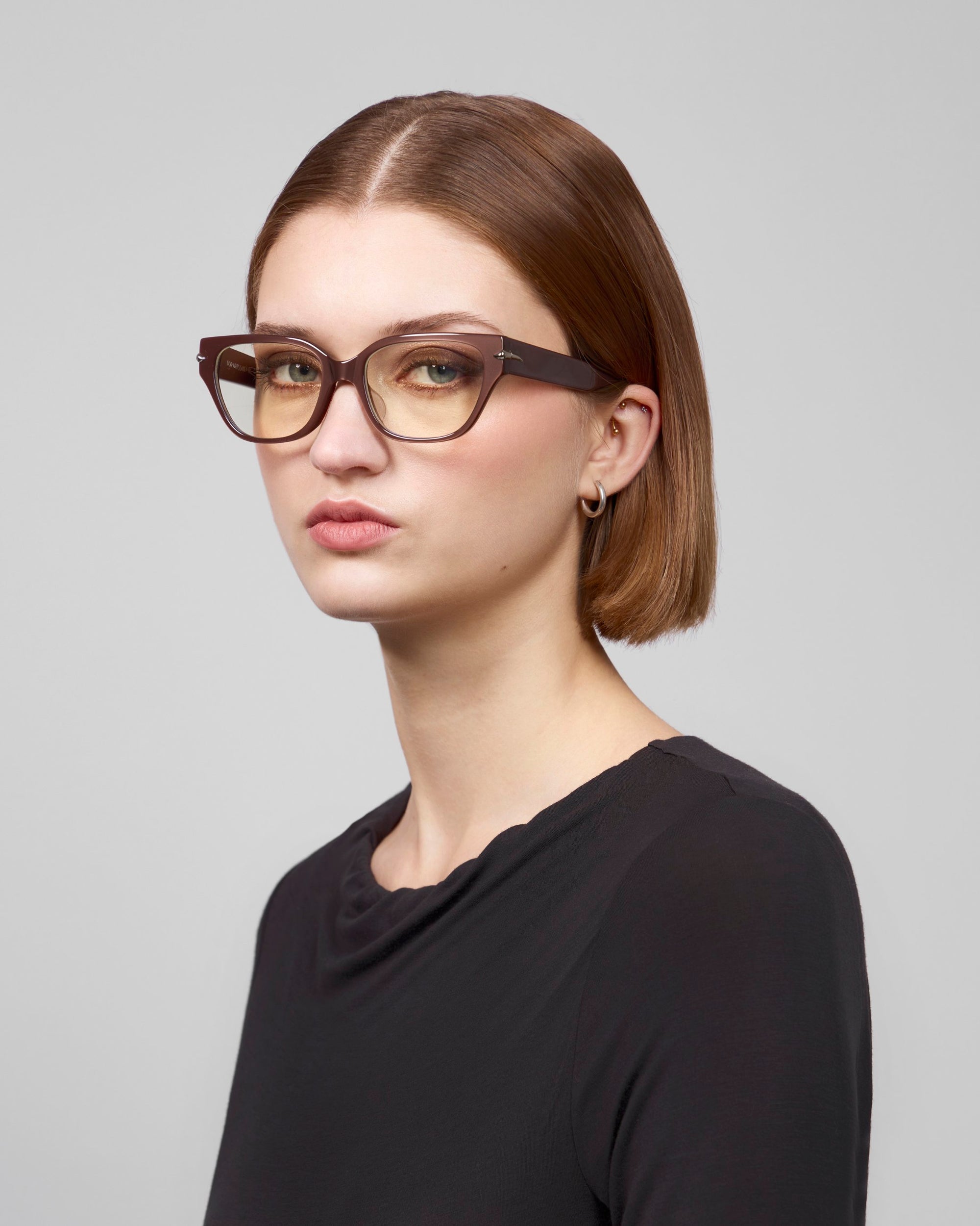 A person with short brown hair wearing Eros black cat-eye acetate frame glasses by For Art's Sake®, along with a black shirt, looks at the camera against a plain, light gray background.