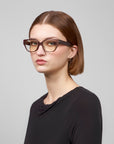 A person with short brown hair wearing Eros black cat-eye acetate frame glasses by For Art's Sake®, along with a black shirt, looks at the camera against a plain, light gray background.