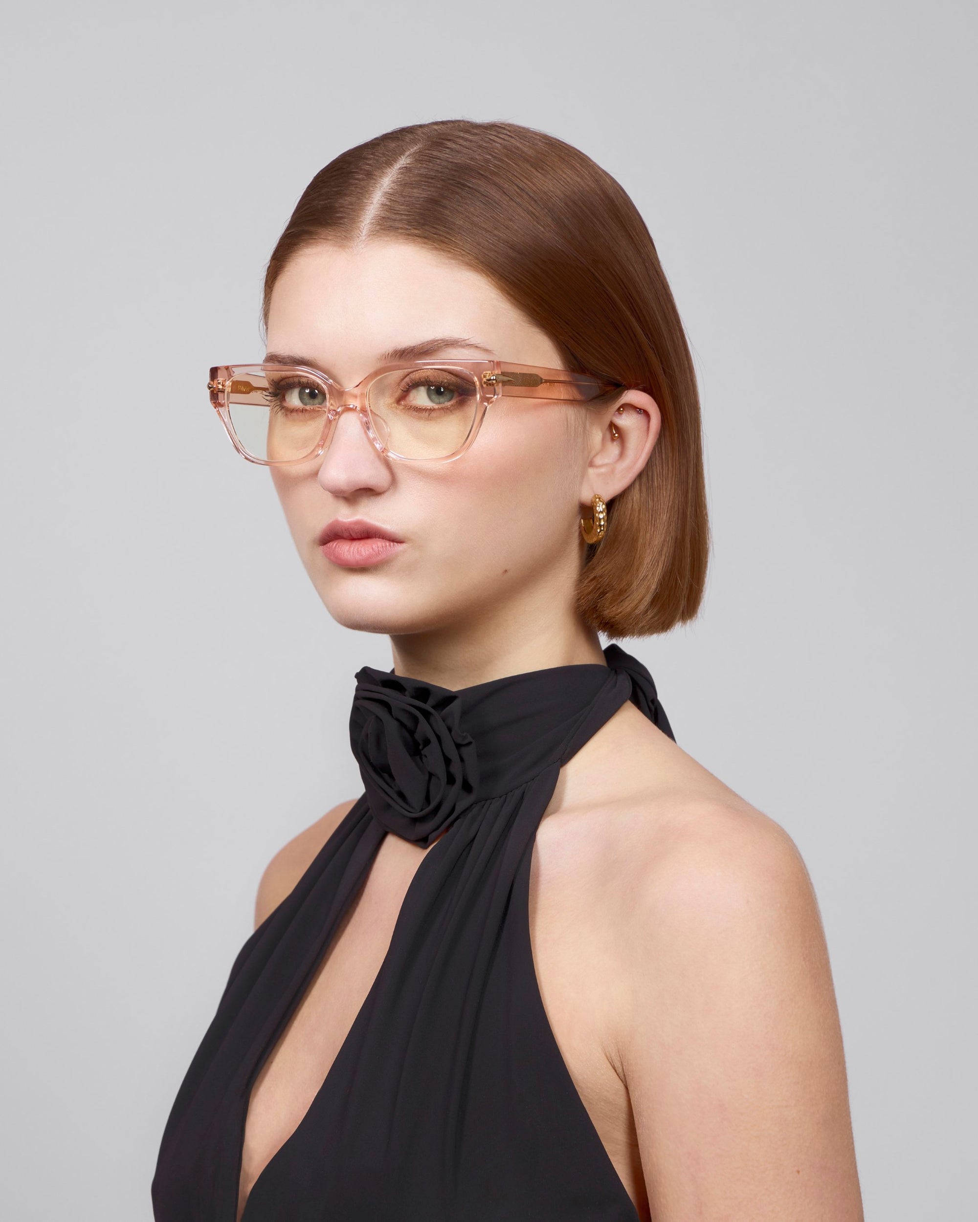 A woman with straight brown hair wears For Art's Sake® Eros cat-eye acetate glasses, gold hoop earrings, and a sleeveless black top featuring a flower detail. She stands against a gray background, looking slightly to the side.