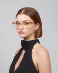 A woman with straight brown hair wears For Art's Sake® Eros cat-eye acetate glasses, gold hoop earrings, and a sleeveless black top featuring a flower detail. She stands against a gray background, looking slightly to the side.