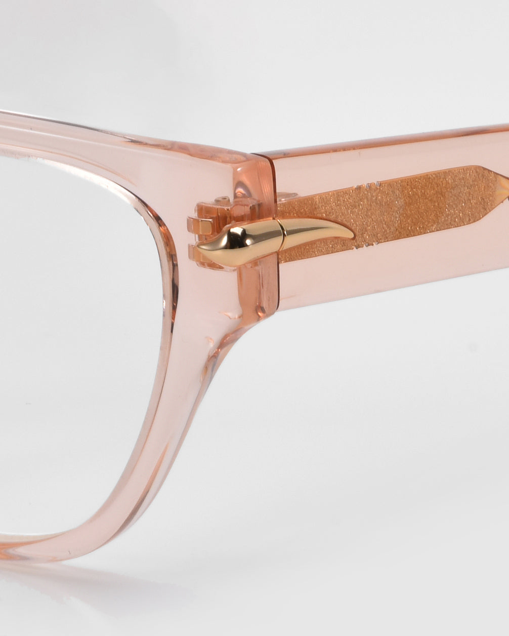 Close-up of Eros by For Art's Sake®: translucent cat-eye acetate frames in light pink, featuring gold metallic horn detail on the hinges. Ideal for everyday wear, these sunglasses add elegance and perfectly complement a rose garden backdrop.