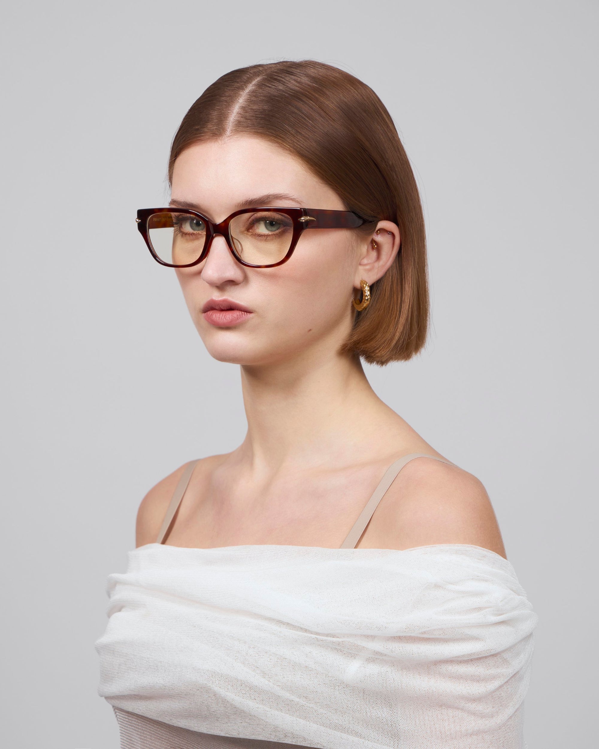 A person with short brown hair, wearing Eros by For Art's Sake® cat-eye acetate frame glasses and gold hoop earrings, gazes at the camera. They wear a white off-shoulder top against a grey background, capturing a refined elegance reminiscent of the Rose Garden collection.