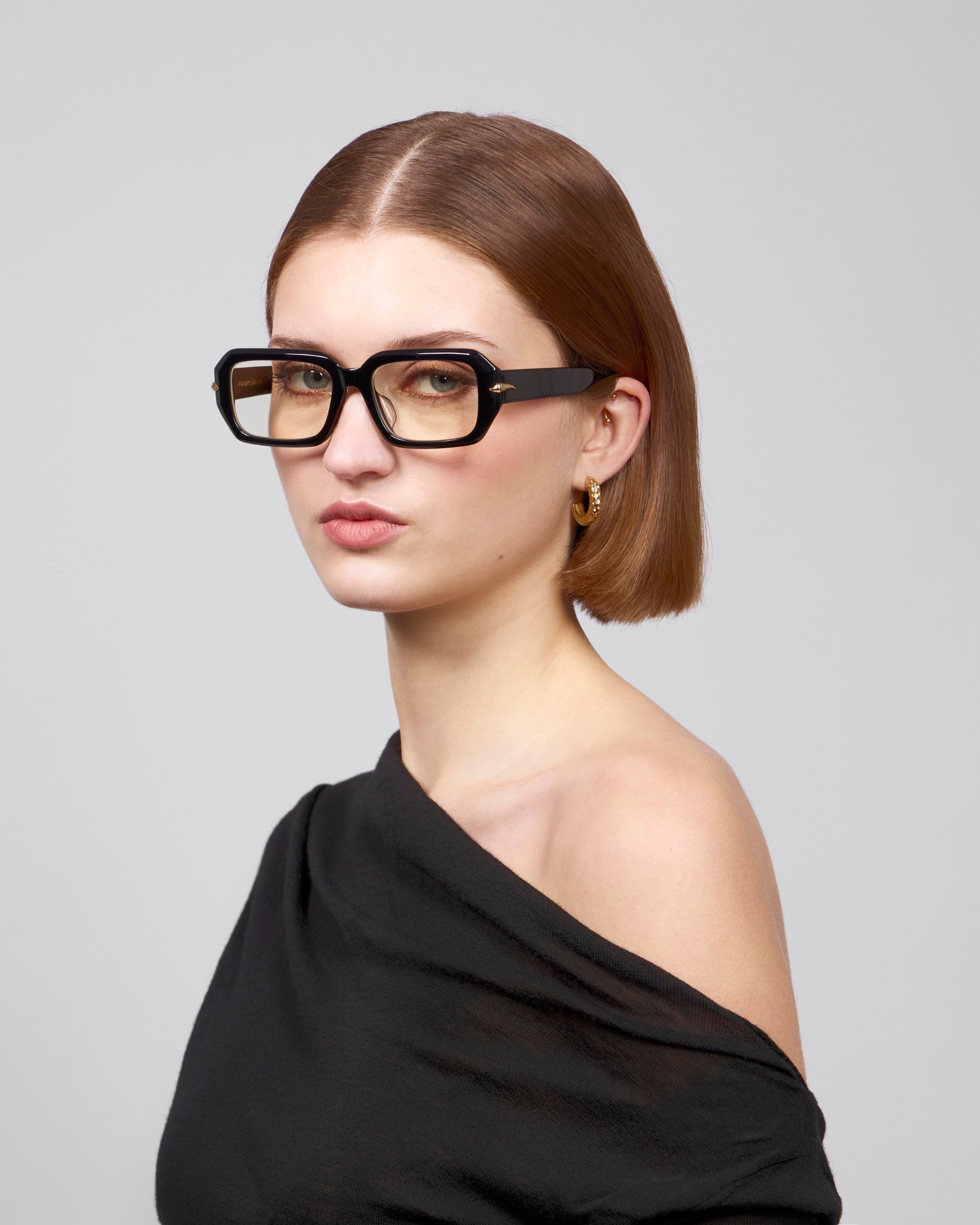 A person with straight brown hair and glasses from the Rose Garden collection wears For Art's Sake®'s Petal top, looking at the camera. The plain gray background subtly contrasts their stylish look.