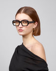 A person with straight brown hair and glasses from the Rose Garden collection wears For Art's Sake®'s Petal top, looking at the camera. The plain gray background subtly contrasts their stylish look.