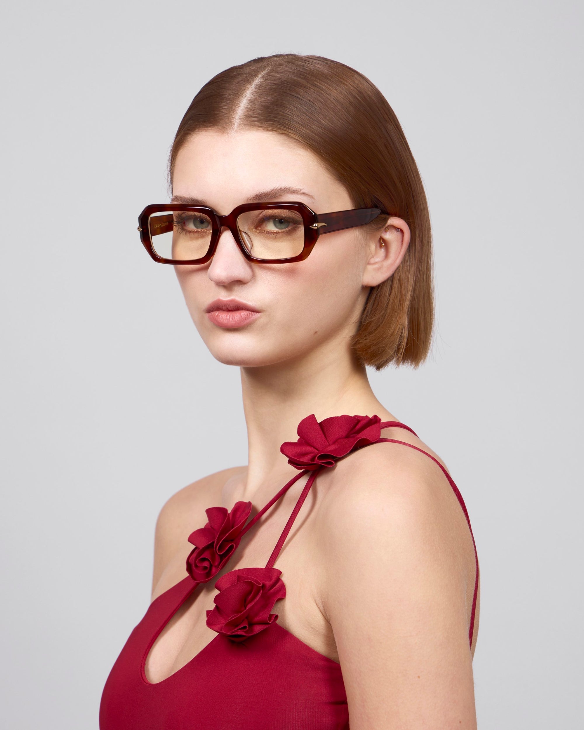 A person with short brown hair wears large, square acetate frames by For Art's Sake® and a red 'Petal' dress with floral strap details, evoking a Rose Garden feel. The background is plain light gray.