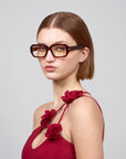 A person with short brown hair wears large, square acetate frames by For Art's Sake® and a red 'Petal' dress with floral strap details, evoking a Rose Garden feel. The background is plain light gray.