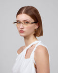 A person with short brown hair and rimless lenses from the Hampton collection by For Art's Sake® gazes neutrally at the camera. They wear a white sleeveless top and golden hoop earrings, set against a plain gray background evoking calming temple decor.