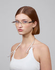 A person with straight, shoulder-length brown hair wears a white sleeveless top and For Art's Sake® Hampton rimless lenses. The background is plain gray, and they have a neutral expression while looking at the camera.