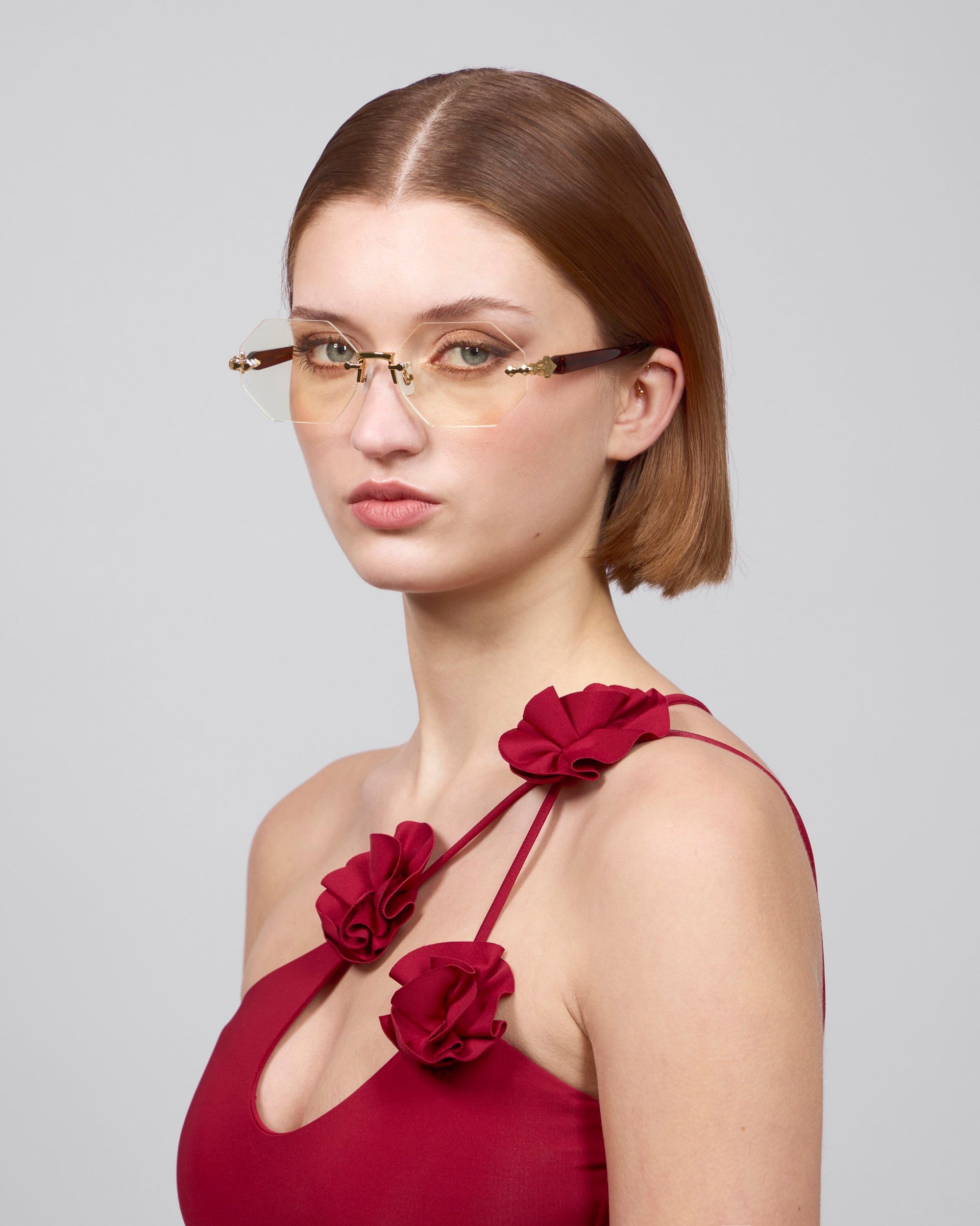 A short-haired person wearing rimless lenses is dressed in the red sleeveless &quot;Hampton&quot; dress by For Art&#39;s Sake®, featuring red fabric flowers on the straps, posing against a plain gray background.