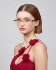 A short-haired person wearing rimless lenses is dressed in the red sleeveless "Hampton" dress by For Art's Sake®, featuring red fabric flowers on the straps, posing against a plain gray background.