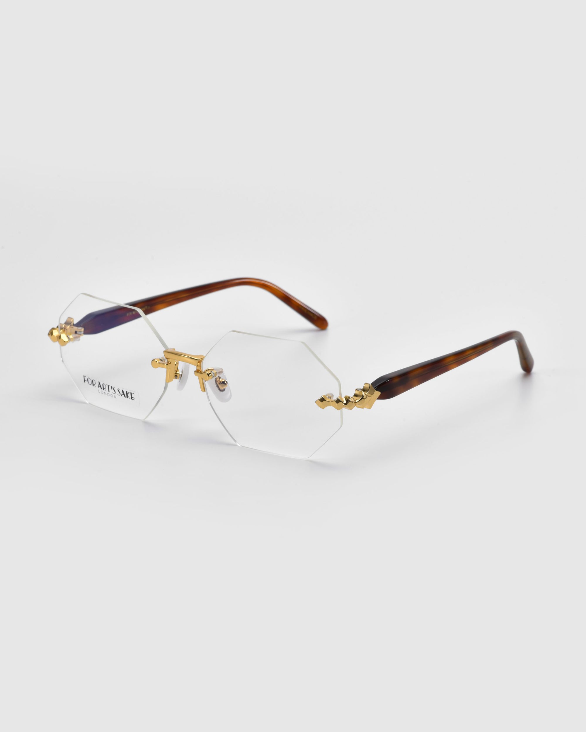 Part of the Rose Garden collection, the &quot;Hampton&quot; rimless glasses from For Art&#39;s Sake® feature hexagonal lenses with gold detailing and dark brown arms. They are showcased on a clean, light gray background that highlights their sophisticated design.