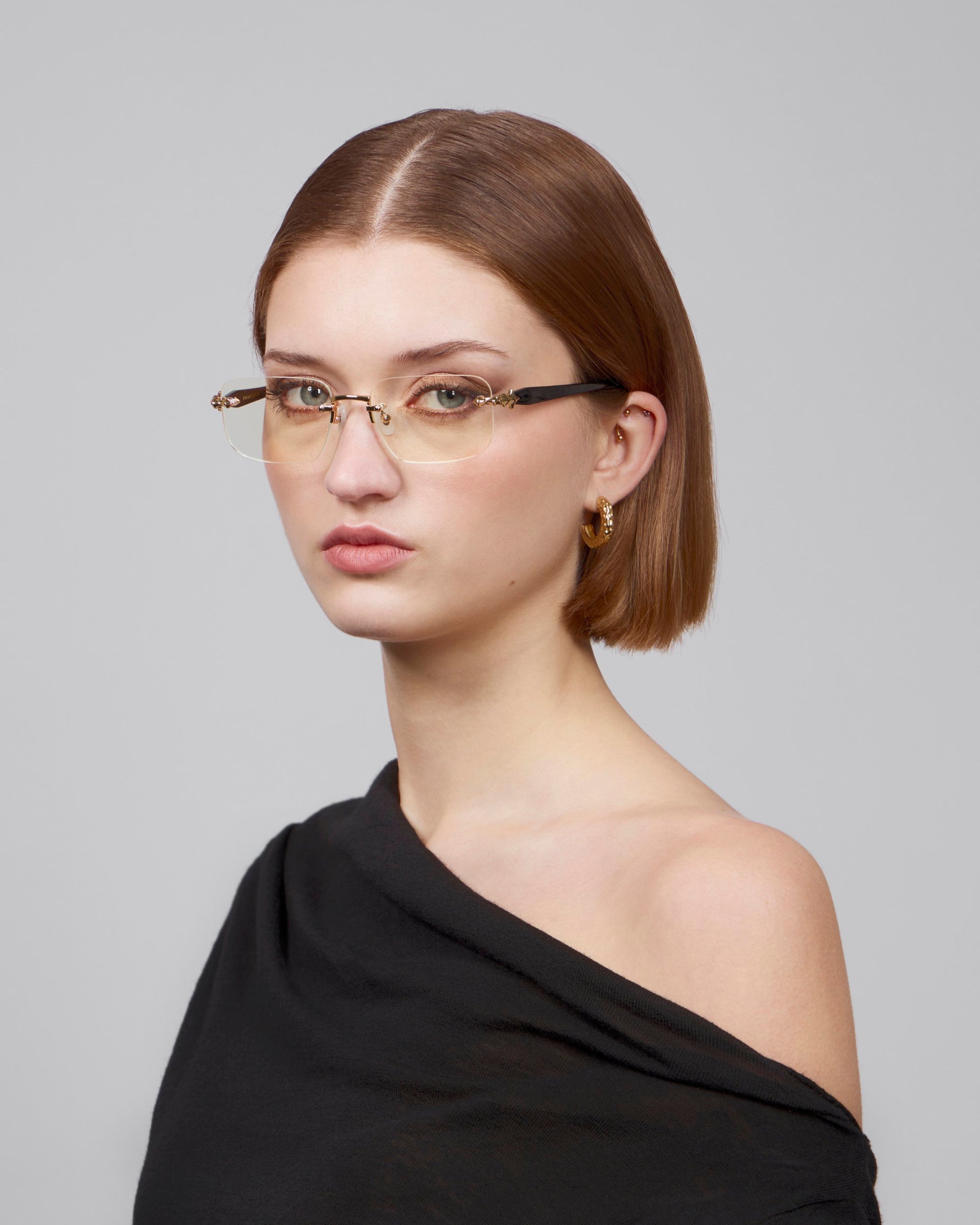 A person with shoulder-length brown hair wears Claremont, thin-rimmed soft rectangular lenses by For Art's Sake®, and a black off-the-shoulder top, creating a timeless classic look against a plain gray background.