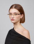 A person with shoulder-length brown hair wears Claremont, thin-rimmed soft rectangular lenses by For Art's Sake®, and a black off-the-shoulder top, creating a timeless classic look against a plain gray background.