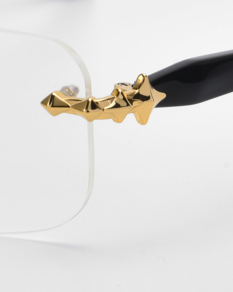 Close-up of the Claremont eyeglasses by For Art's Sake® from the Rose Garden collection, showcasing soft rectangular lenses. The transparent frame pairs with black temple arms embellished with gold stars near the hinge, set against a simple white background to emphasize their elegance.