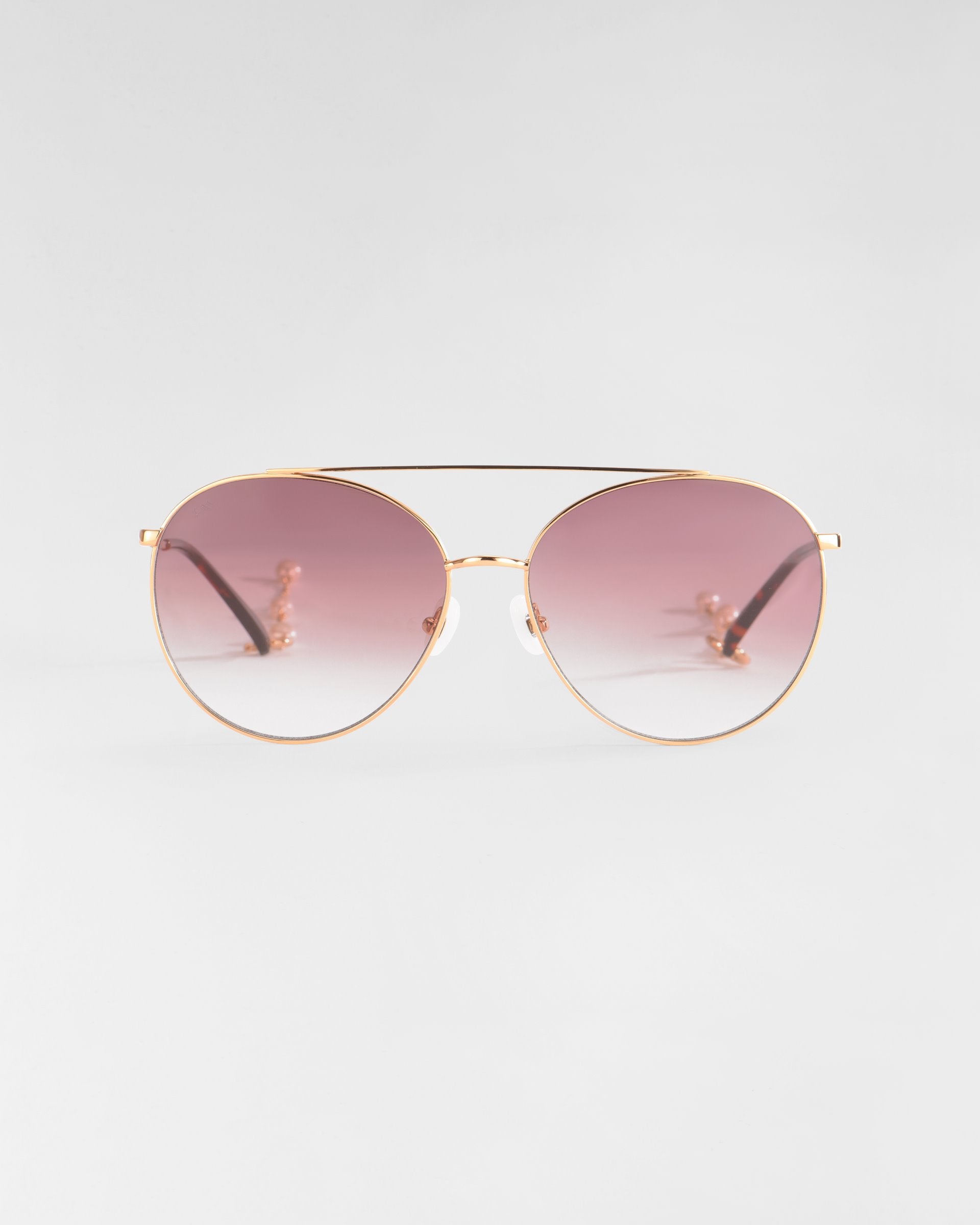 A pair of stylish aviator sunglasses named Yoyo by For Art's Sake® with 18k gold-plated metal frames and gradient pink-tinted lenses is centered against a plain, light gray background. The Yoyo sunglasses feature thin arms and adjustable nose pads for comfort.