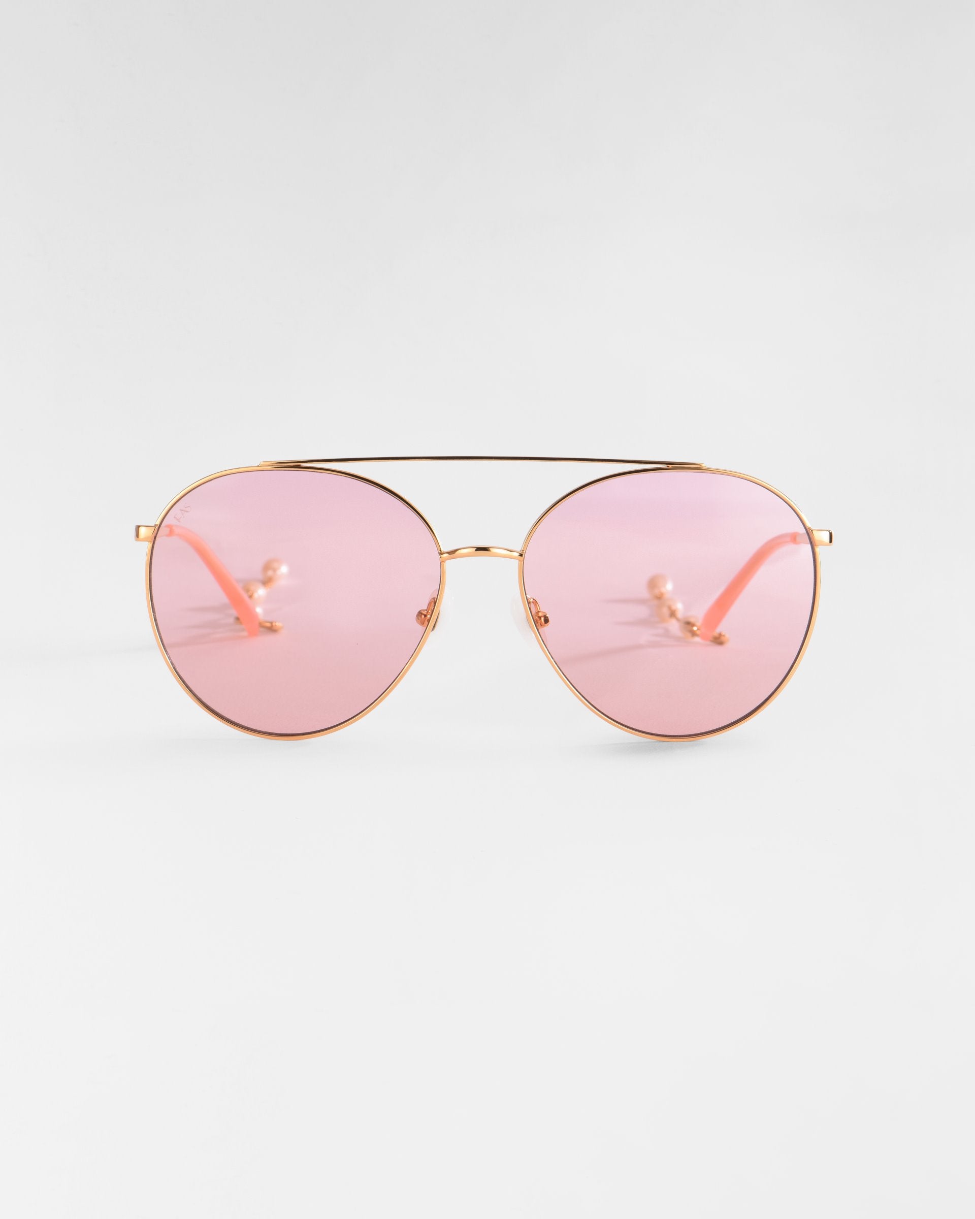 A pair of fashionable aviator sunglasses featuring 18k gold plating on the frame and light pink tinted lenses. The bridge and nose pads are also gold, with the stems extending to slender, light pink ear pieces. The background is a plain, neutral gray. This is the Yoyo by For Art's Sake®.