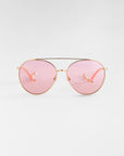 A pair of fashionable aviator sunglasses featuring 18k gold plating on the frame and light pink tinted lenses. The bridge and nose pads are also gold, with the stems extending to slender, light pink ear pieces. The background is a plain, neutral gray. This is the Yoyo by For Art's Sake®.