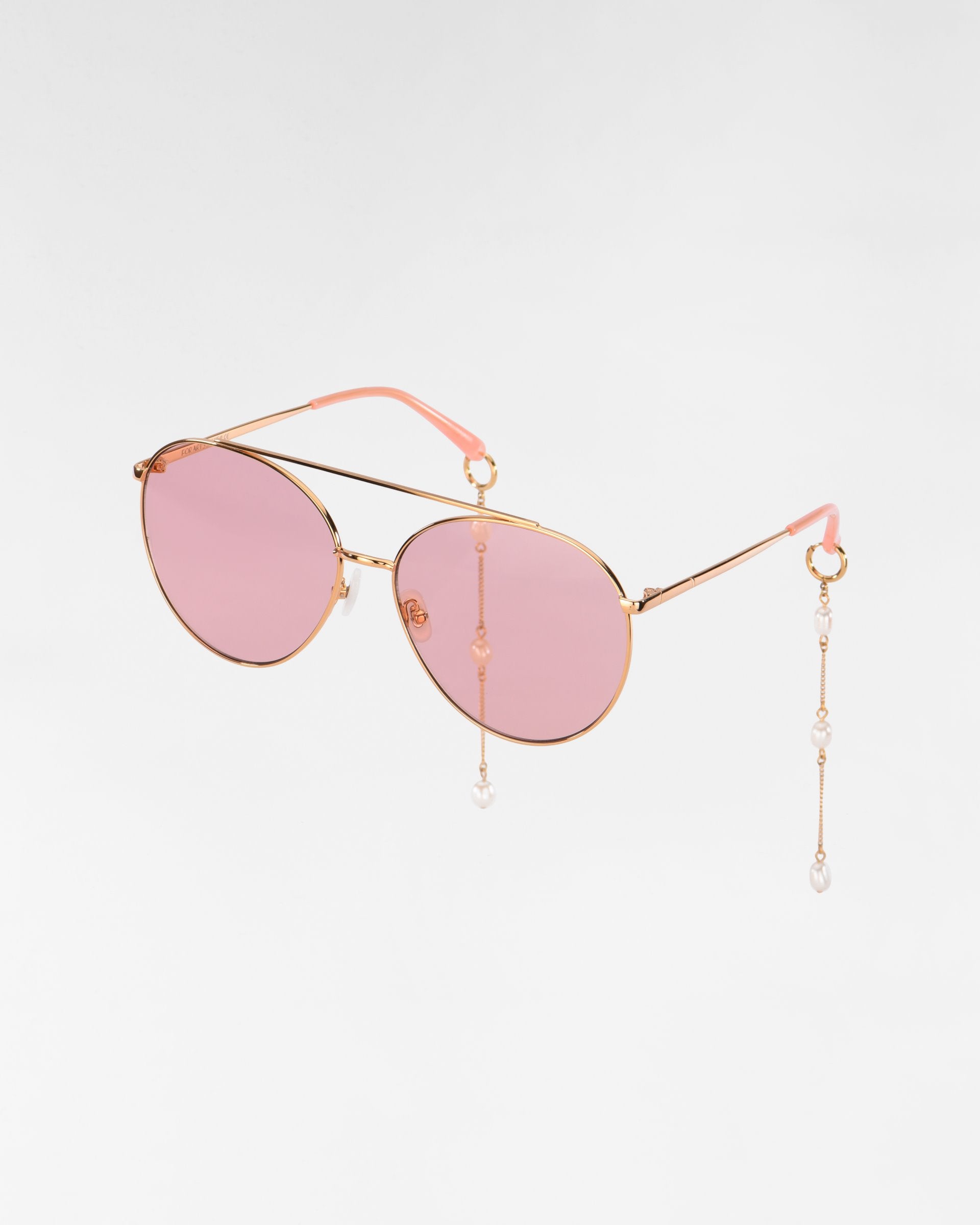 Yoyo by For Art&#39;s Sake®: Gold-framed aviator sunglasses with pink-tinted lenses, adorned with delicate freshwater pearl-chains hanging from the arms. The sunglasses have pink accents at the ends of the arms, adding a touch of elegance and style. Featuring 18k gold plating for an extra luxurious finish, against a plain white background.