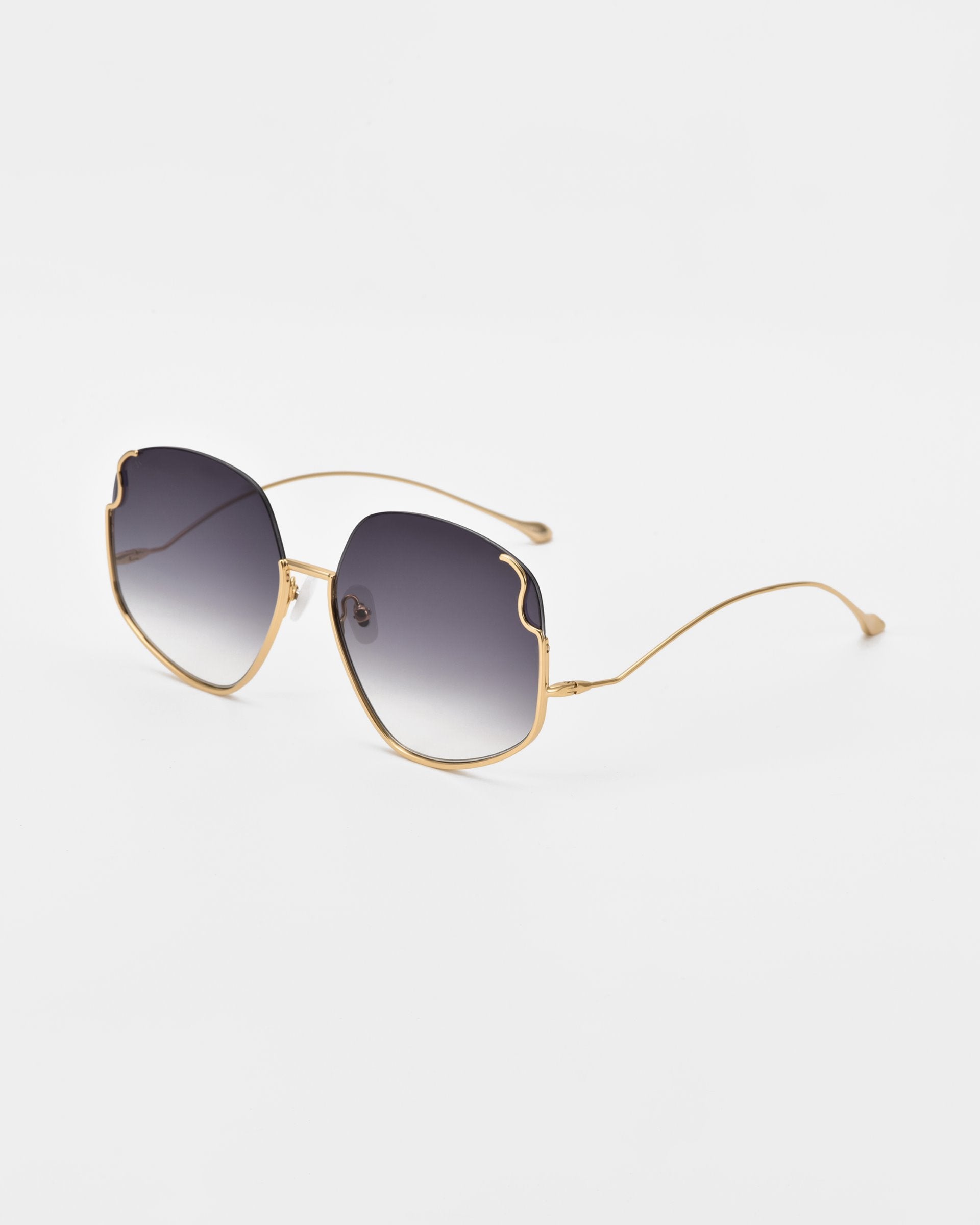 A pair of luxurious For Art&#39;s Sake® Drape sunglasses with gold metal frames and dark gradient lenses. The frames feature intricate metal detailing and a unique, slightly irregular shape at the top edges. The elegant arms are thin and curve towards the back ends, where there are small, rounded tips. These sunglasses are set against a plain white background.
