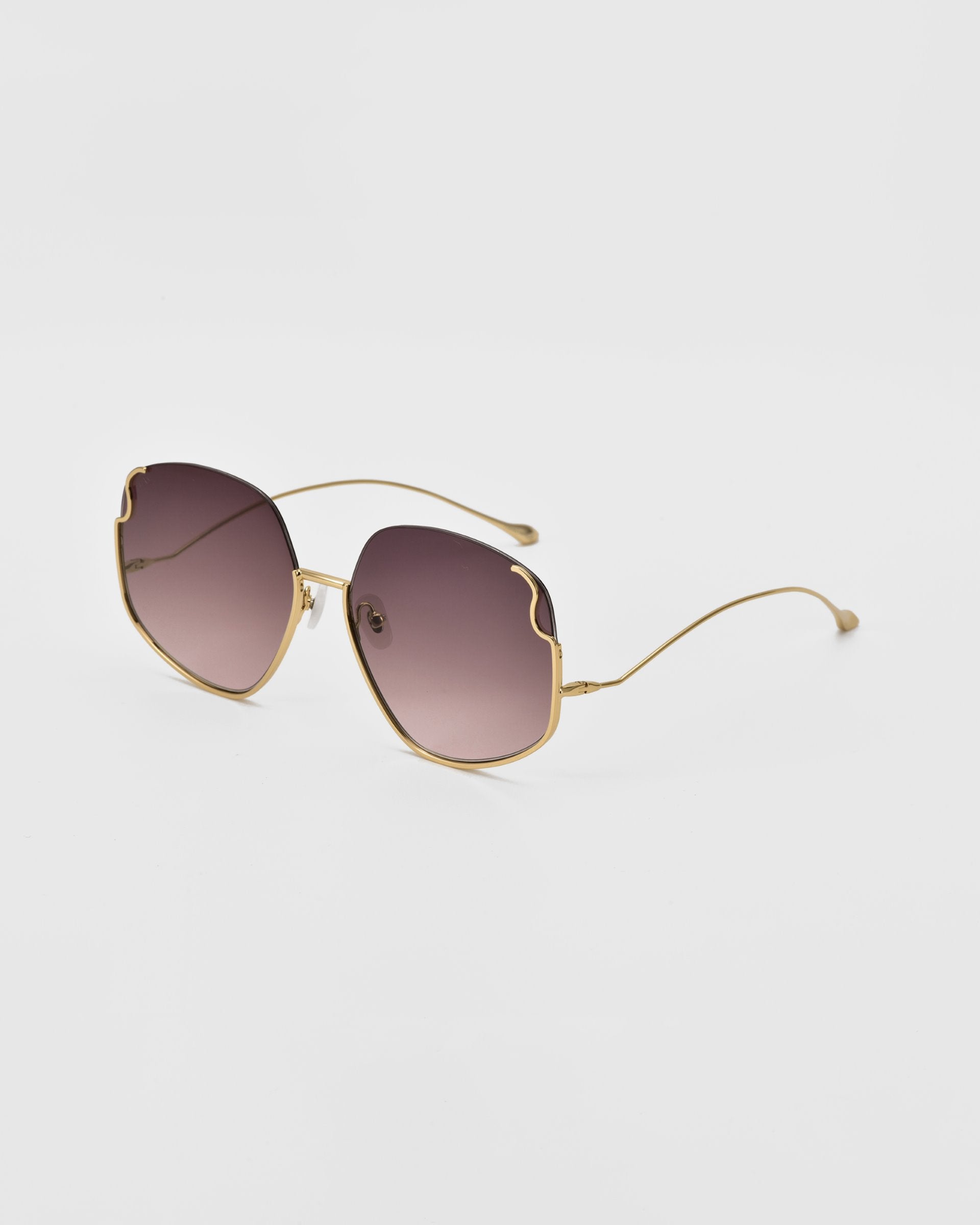 A pair of Drape sunglasses by For Art&#39;s Sake® with gold metal frames and gradient lenses. The oversized squared frame transitions from dark at the top to lighter at the bottom. These sunglasses feature a unique curved design on the upper corners of the lenses, complete with intricate metal detailing.