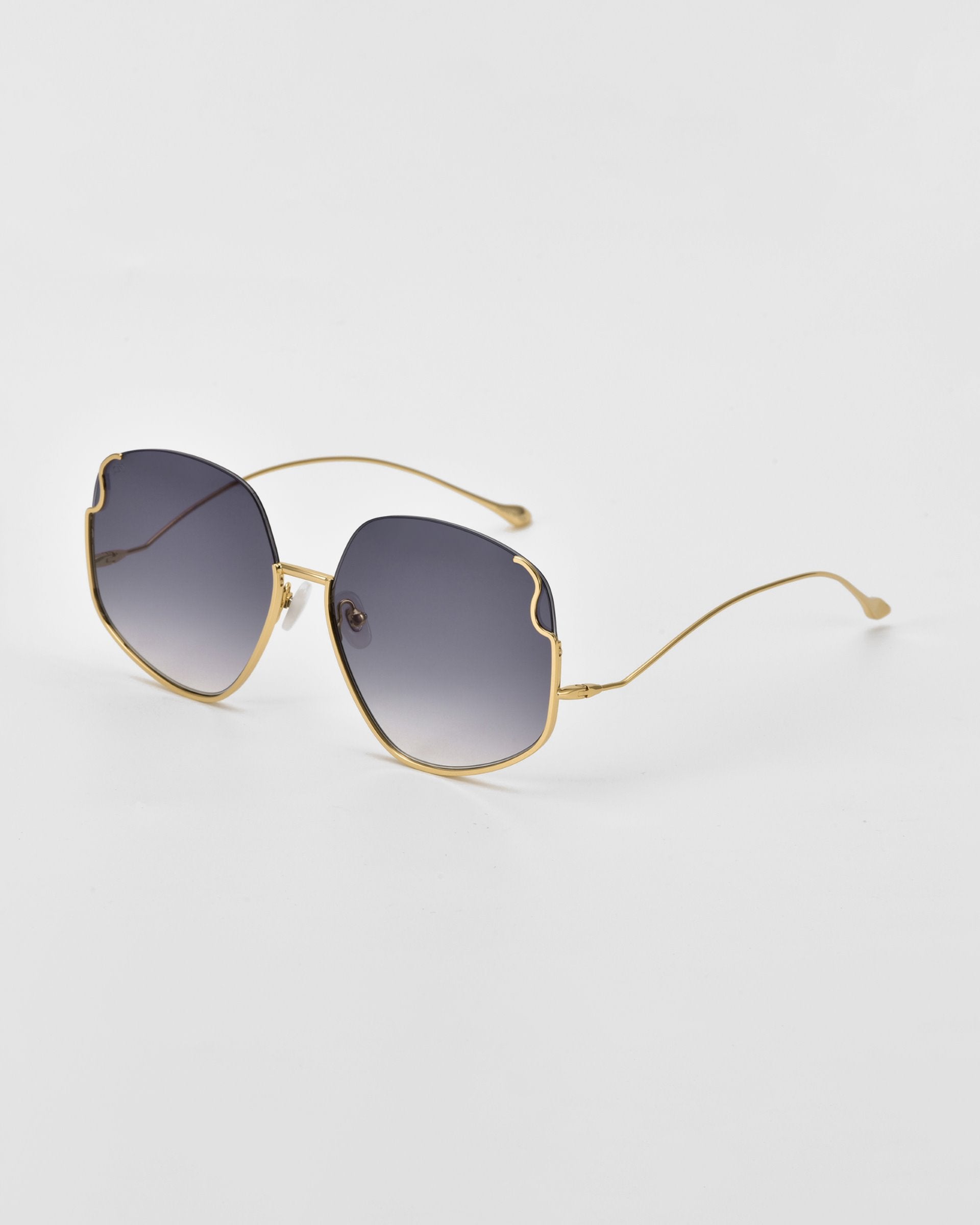 A pair of Drape sunglasses by For Art&#39;s Sake® with gold frames and dark, gradient lenses. The design features a unique, slightly wavy cut on the top edges of the oversized squared frame. Intricate metal detailing adds to their charm as they rest on a white surface, viewed at an angle.